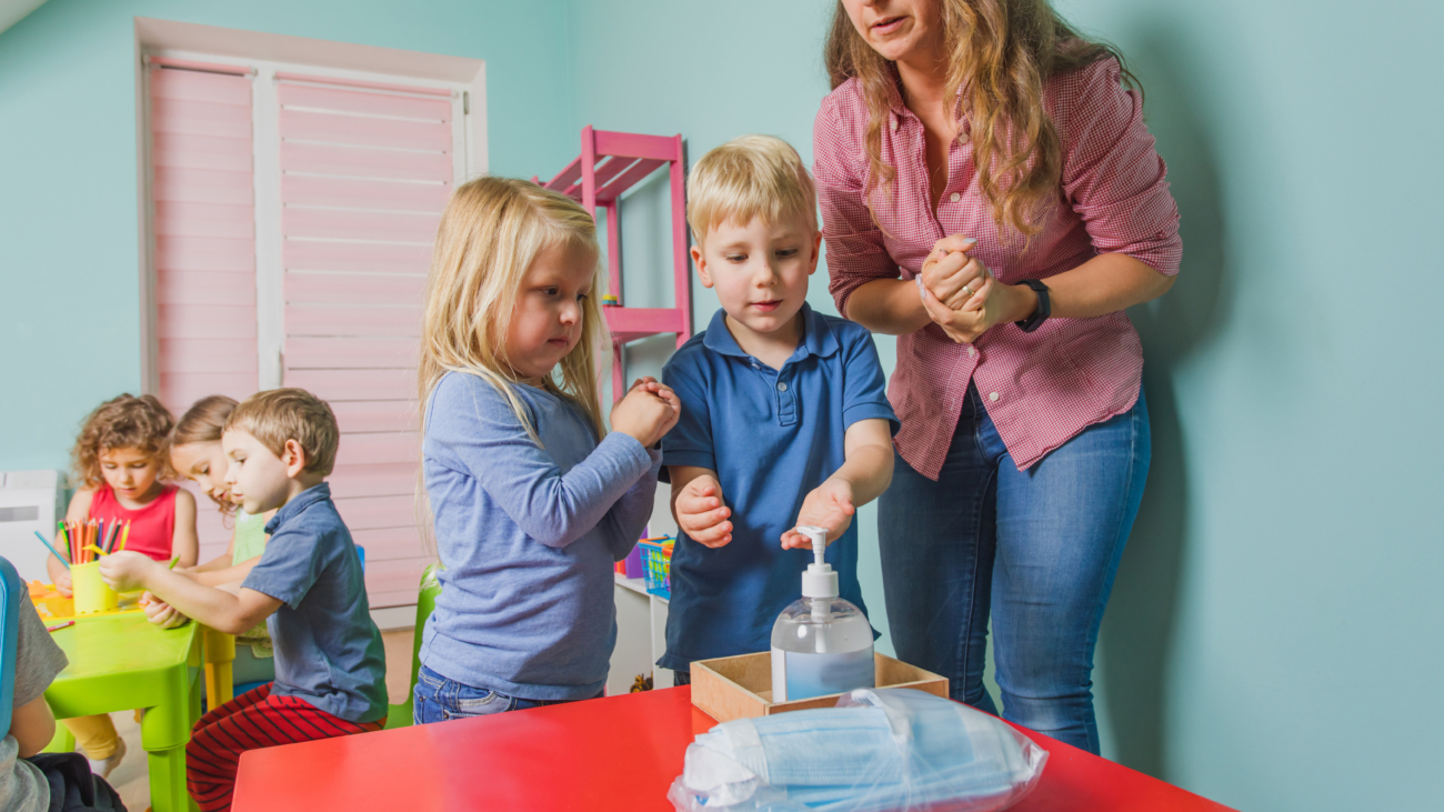 Young teacher at the preschool shows children how to use sanitazer and clean their hands. New hygienic rules at the kindergartens and schools to teach children to protect their health. New normal.