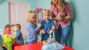 Young teacher at the preschool shows children how to use sanitazer and clean their hands. New hygienic rules at the kindergartens and schools to teach children to protect their health. New normal.
